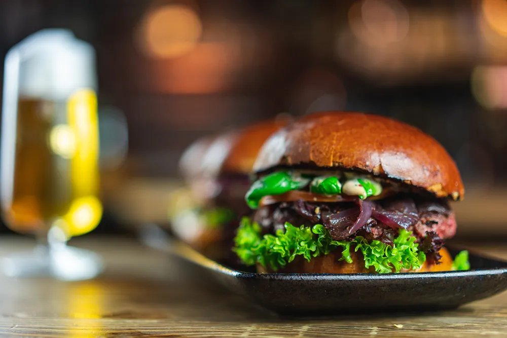 Broderick Burger mit karamellisierten Zwiebeln, Tomate und Basilikum, dahinter ein frisch gezapftes Bier